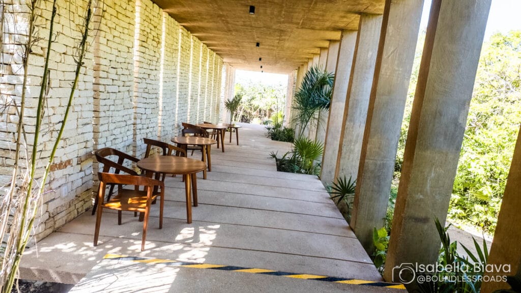 A serene outdoor corridor features wooden tables, chairs, stone walls, and lush greenery, creating a peaceful setting under a partially enclosed concrete roof.