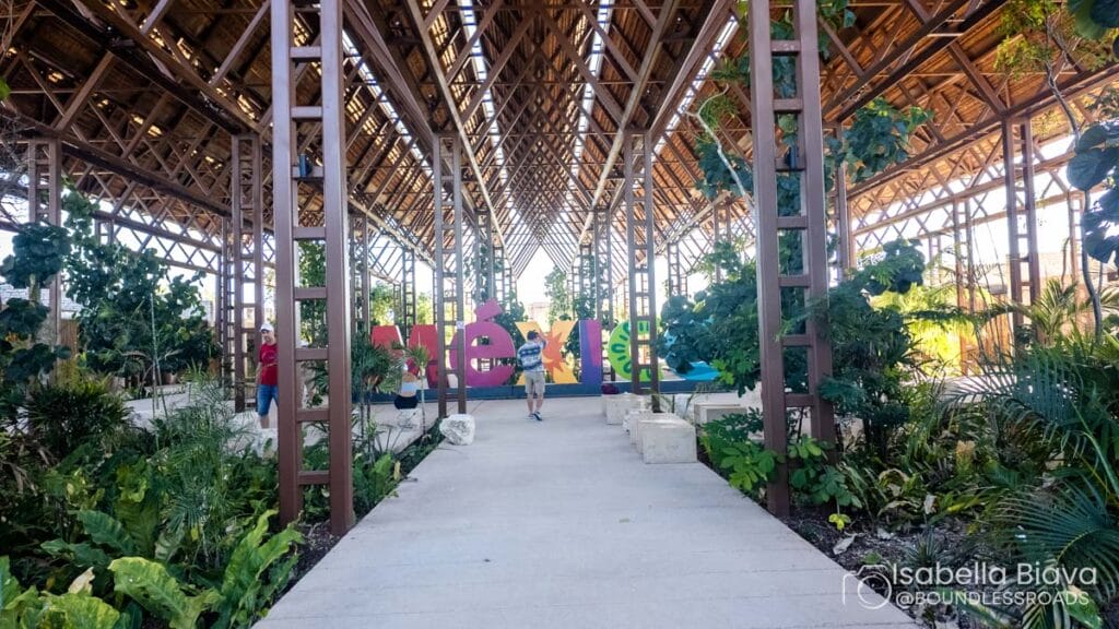 A modern structure with open beams features vibrant "MÉRIDA" letters, surrounded by lush greenery and several people walking around.