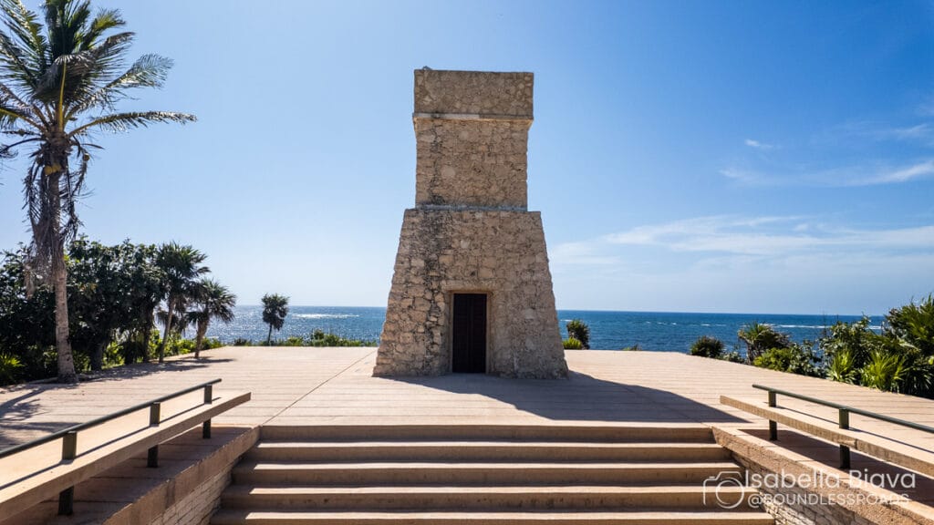 Stone structure by the ocean in Tulum, surrounded by palm trees. Clear blue sky and water with stairs leading up.