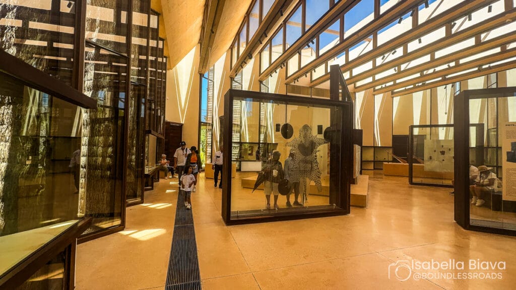 Modern museum interior with geometric architecture, natural light streaming through windows. Several people, including children, explore exhibits in glass cases. Bright, airy atmosphere.