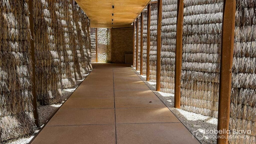 A modern pathway features thatched walls and a wooden ceiling, leading toward a stone wall, bathed in natural sunlight.