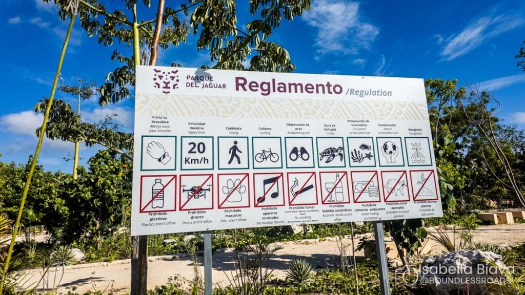 Sign at Parque del Jaguar lists regulations, speed limit, and prohibits activities like smoking and drinking. Surrounded by trees under a blue sky.