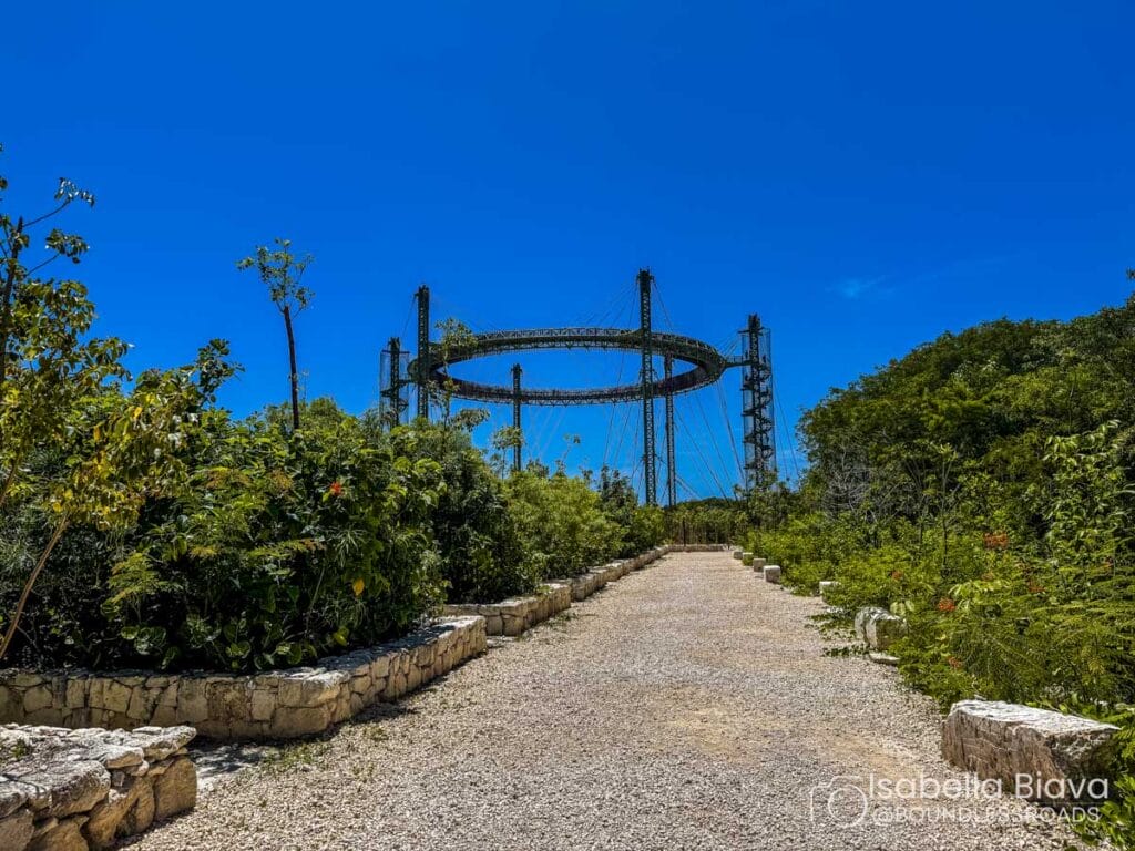 A gravel path leads toward the Orbit Tower at Parque Ecológico Xcaret, surrounded by lush greenery under a clear blue sky.