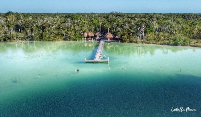 Clear turquoise water surrounds a wooden pier with thatched huts, bordered by lush forest. A person wades through the serene lagoon.
