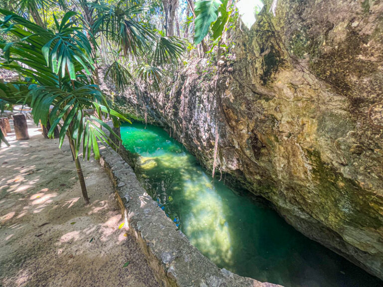 A narrow, turquoise river flows between rocky walls and tropical plants, with sunlight highlighting the natural beauty and lush greenery surrounding the area.