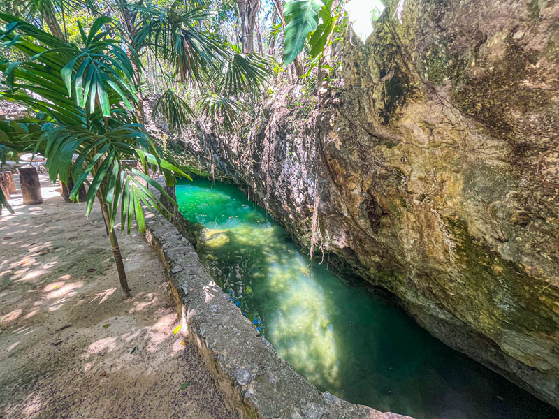 A narrow, turquoise river flows between rocky walls and tropical plants, with sunlight highlighting the natural beauty and lush greenery surrounding the area.