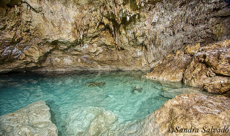 Crystal-clear turquoise water pool inside a cave, surrounded by rocky walls and stalactites, creating a serene and natural underground setting.