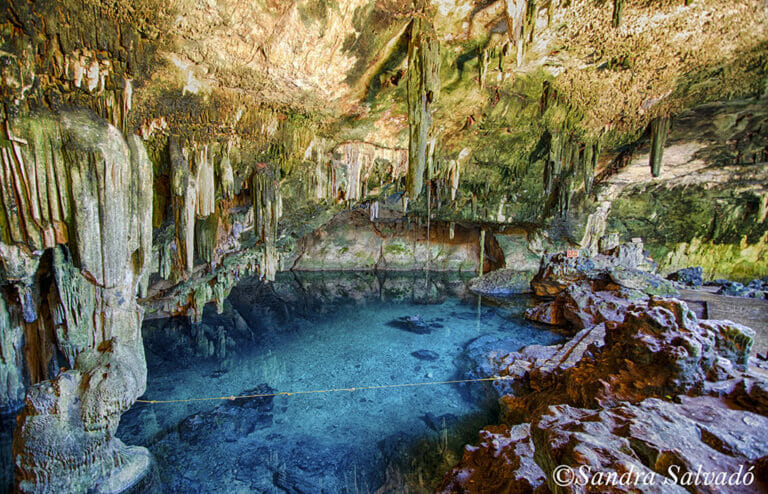 A stunning cave interior with striking stalactites, stalagmites, and clear blue water, creating a serene, otherworldly atmosphere.