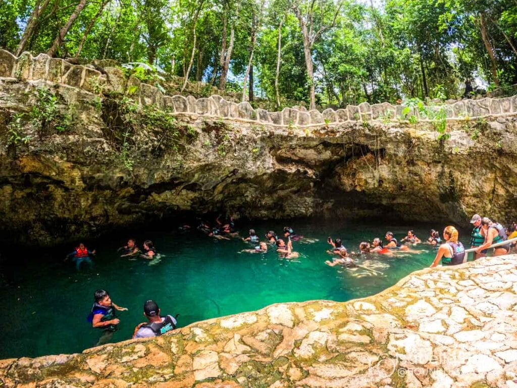People swimming in a natural cenote surrounded by lush greenery and rocky formations, enjoying a tropical outdoor adventure experience.