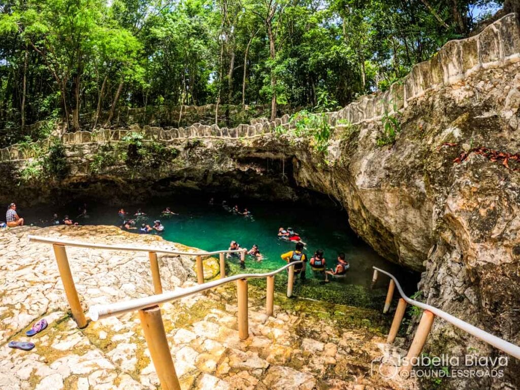People swimming in a natural cenote surrounded by dense forest, accessed by stone steps, creating a picturesque and serene setting.