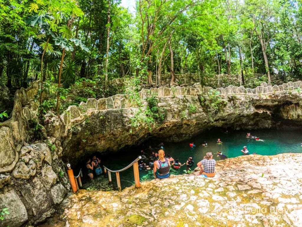 People swim and relax by the edge of a natural cenote surrounded by lush greenery and rocky formations.