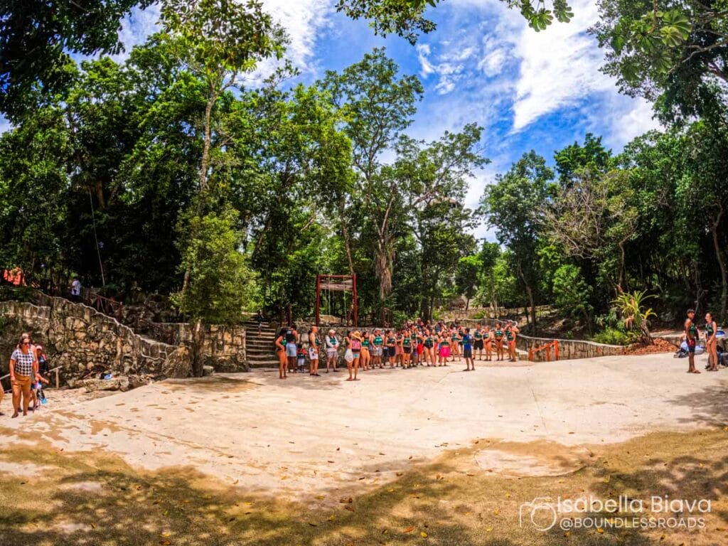 A group of people gathers in a lush, forested area near stone stairs under a partly cloudy sky, creating a vibrant outdoor scene.