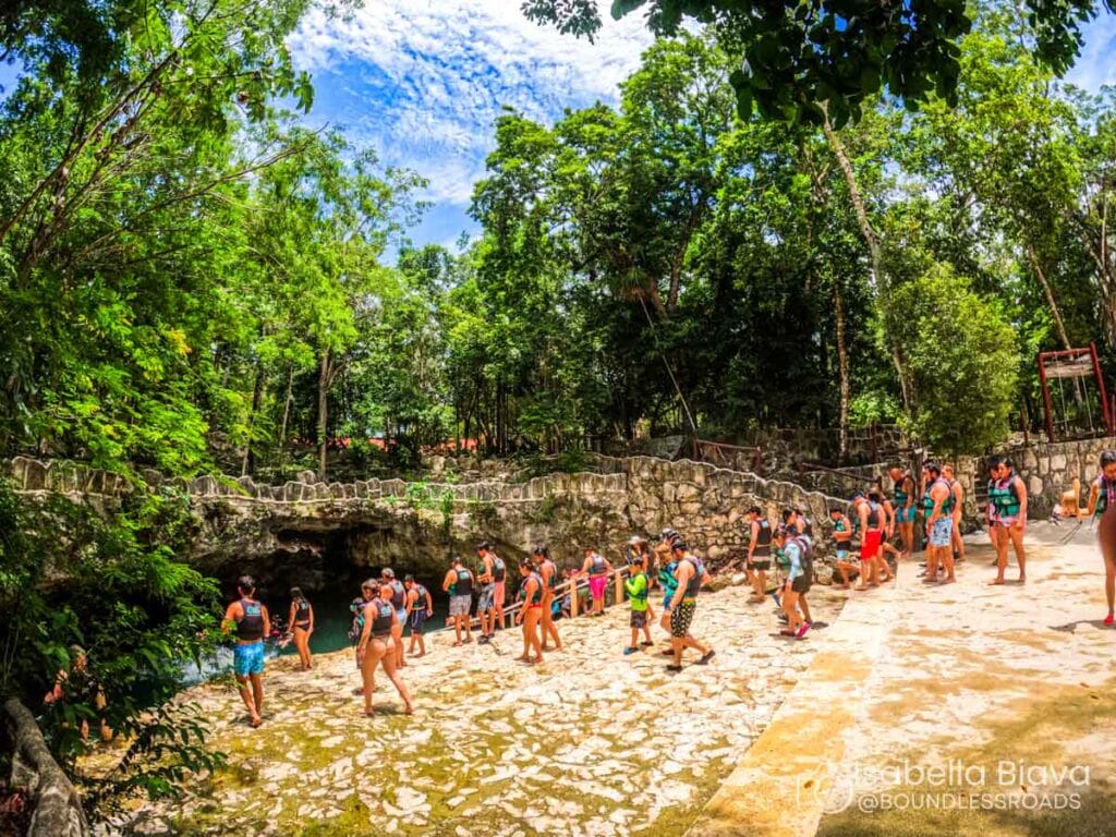 A group of people in swimwear explore a lush, dense forest area with a rocky entrance, possibly a cenote or cave.