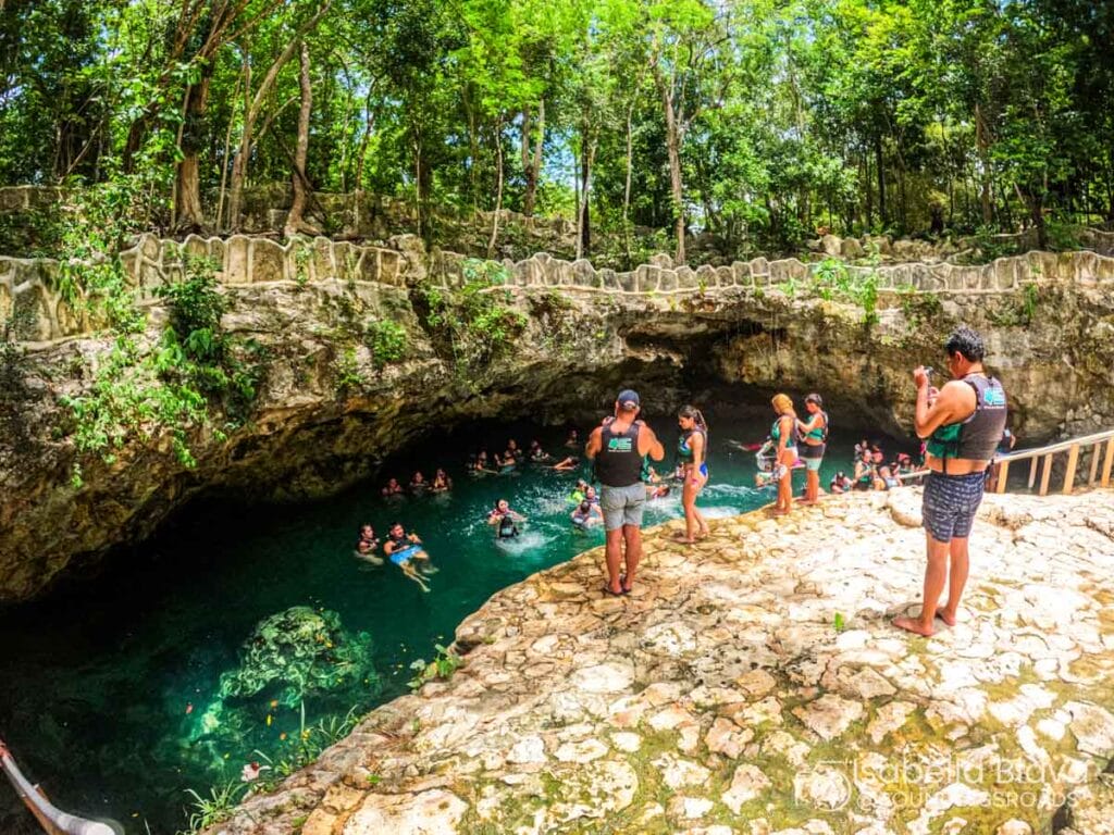 People wearing life vests enjoy swimming in a natural cenote surrounded by lush trees and rock formations, creating a serene, adventurous atmosphere.