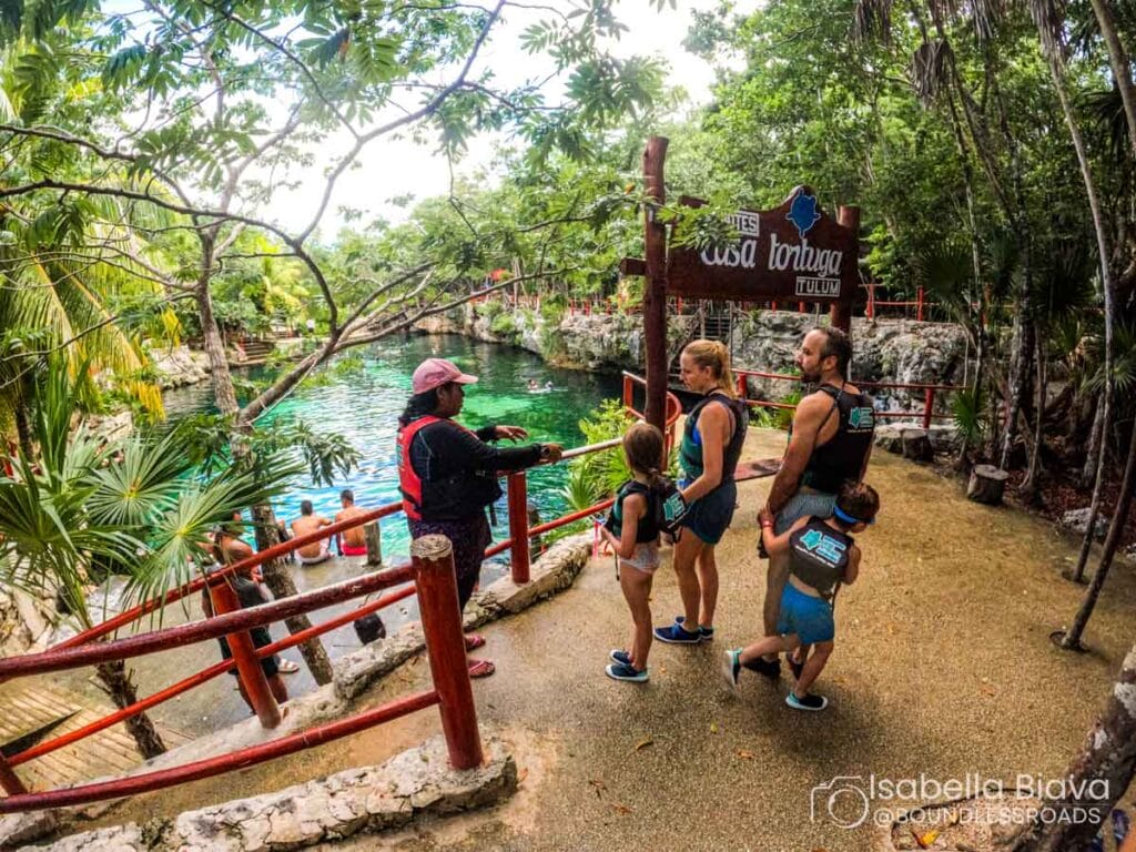 A group of people in life vests stand by a scenic water area labeled Casa Tortuga Tulum, surrounded by lush trees and red railings.