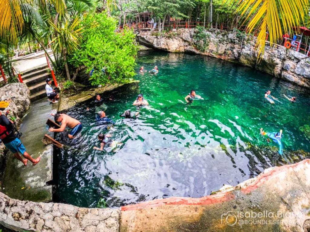 People swimming and relaxing in a natural cenote surrounded by lush greenery, rocky edges, and tropical vegetation, with a vibrant, inviting atmosphere.