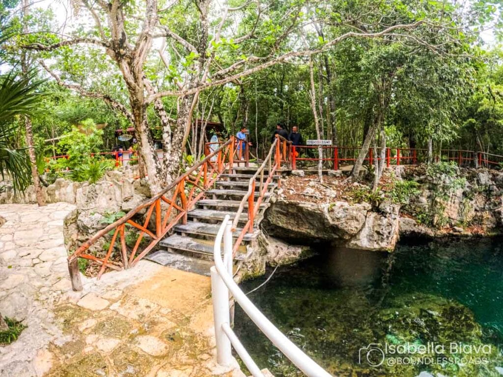 A natural swimming area with a raised wooden walkway, surrounded by lush greenery. People stand on the path, enjoying the scenic view.