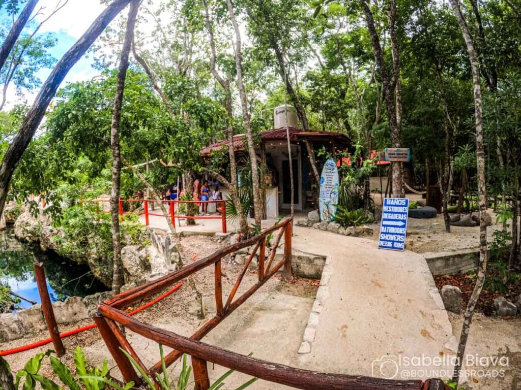 A small entrance with signage and people is surrounded by lush forest and a pathway leading to a natural water area.