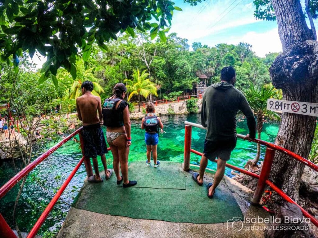 Four people in swimsuits stand on a platform near a tropical, clear blue water cenote, surrounded by lush greenery and vibrant palms.