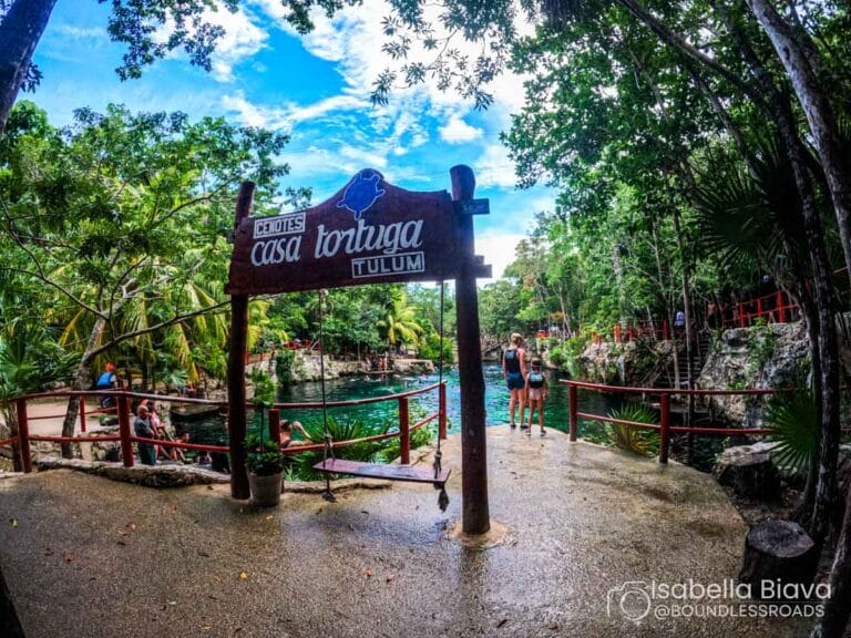 A sign reads "Casa Tortuga Tulum" at a scenic, tropical cenote. People explore the vibrant, lush surroundings under a bright, clear sky.