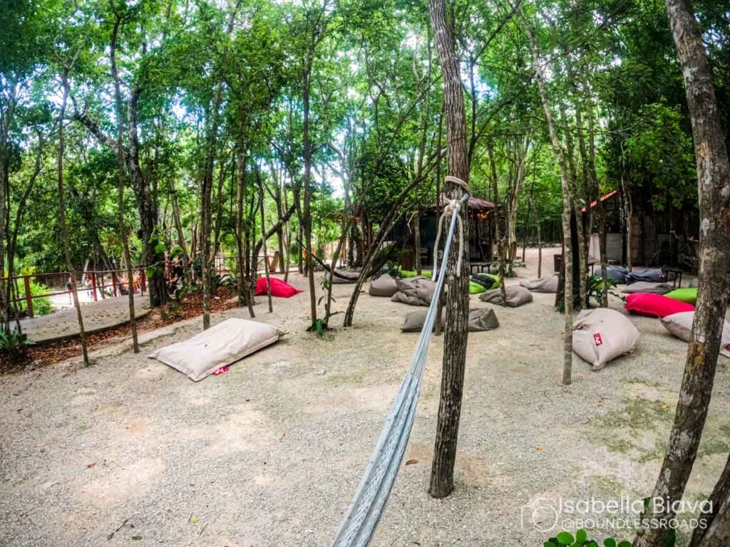 Outdoor relaxation area with hammocks and bean bags among trees. People in the background enjoy the serene, natural setting.
