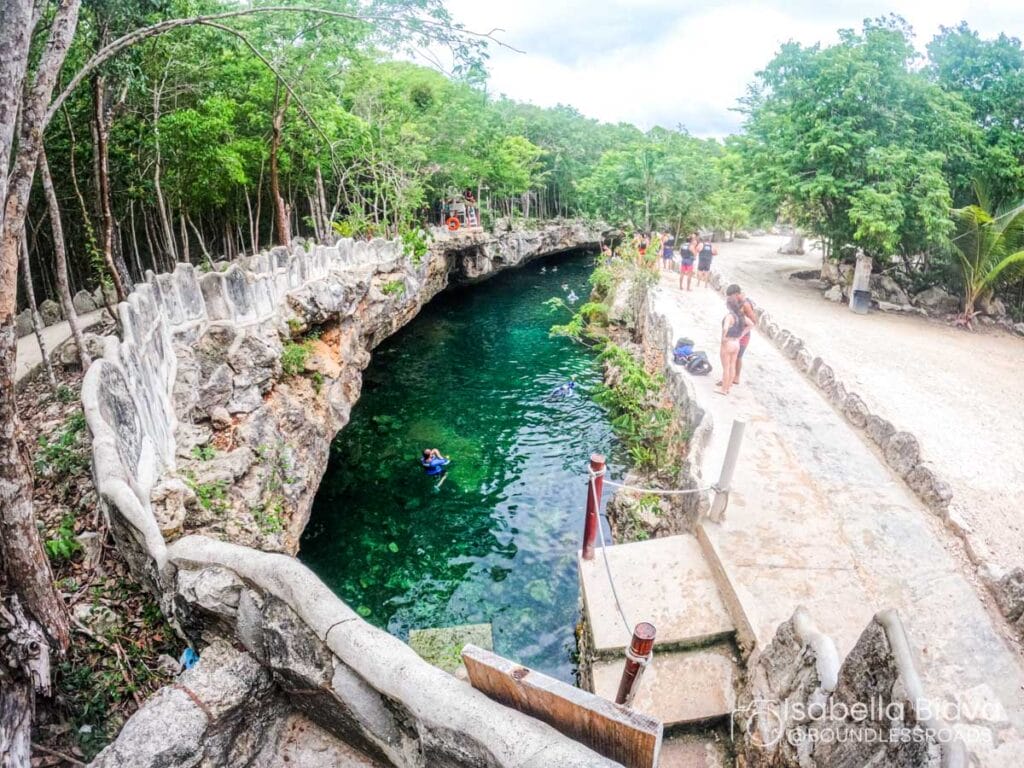 A serene cenote surrounded by rocky cliffs and lush greenery, with people relaxing and swimming in the clear water.