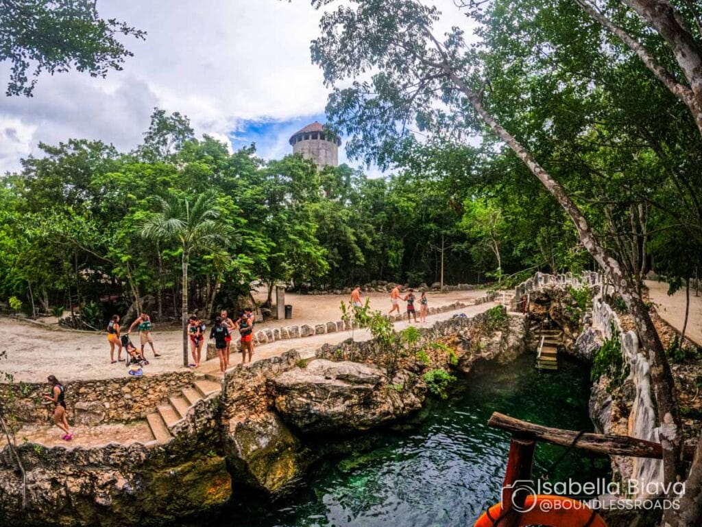 A group of people enjoy a lush, natural setting near a stone structure, with clear water and lush greenery surrounding them.
