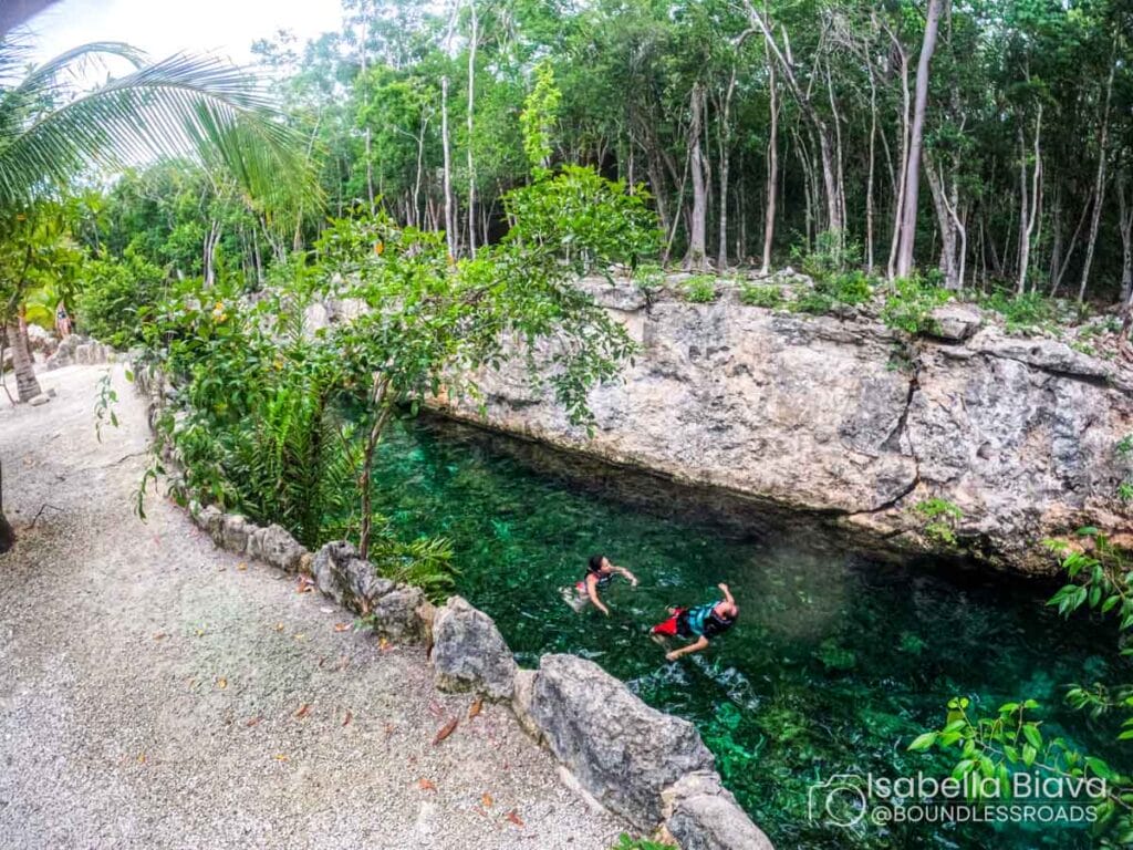 Two people swim in a clear, rocky cenote surrounded by lush greenery and trees, creating a tranquil natural scene.