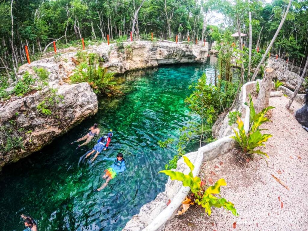 Clear turquoise water cenote surrounded by rock formations and lush greenery. Several persons swim and snorkel, enjoying the natural scenery.
