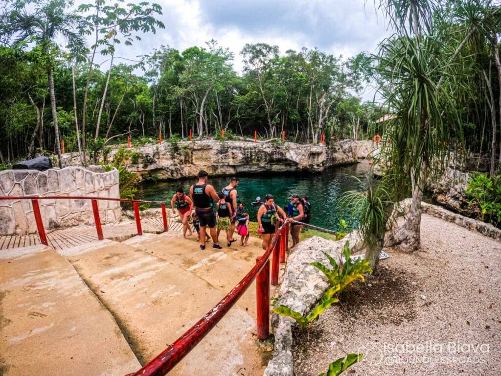 A group of six people prepares to enter a natural cenote surrounded by lush trees and rocky terrain.