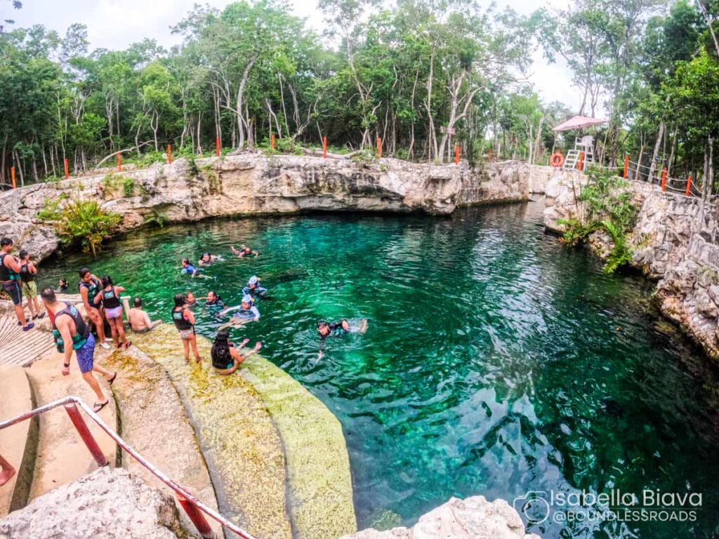 People swim in a natural cenote surrounded by rock formations and trees, with clear turquoise water and safety railings around the edge.