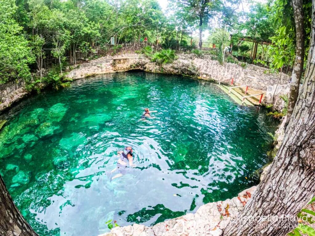 A natural cenote surrounded by lush trees, with two people swimming in the clear turquoise water near stone steps and walls.
