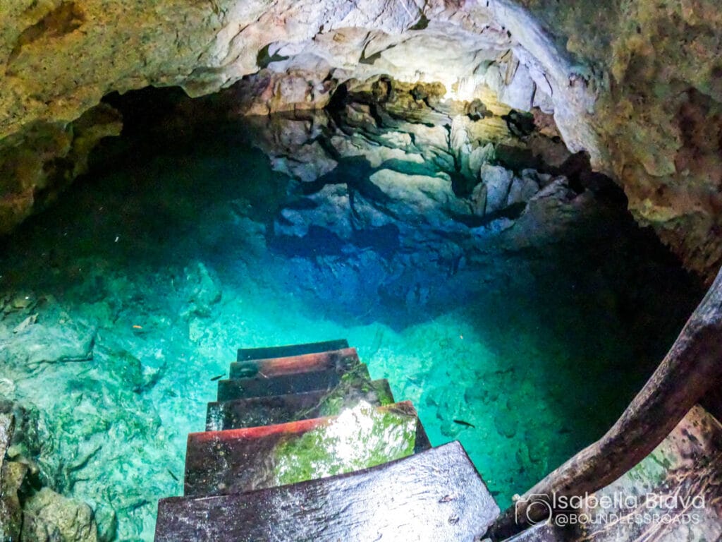 Stairs descend into a turquoise water-filled cave with rocky walls, illuminated by natural light, creating a serene, mesmerizing underground scene.