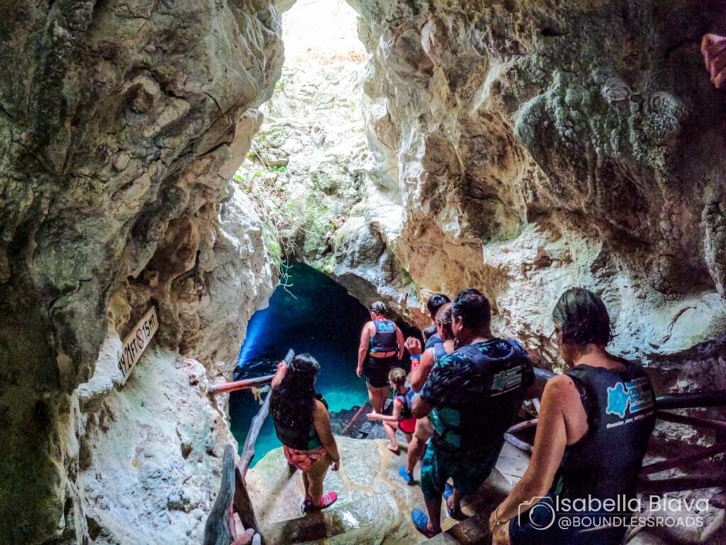 People wearing life vests descend stairs into a rocky cave with clear blue water. It's a natural swimming spot inside a cavern.