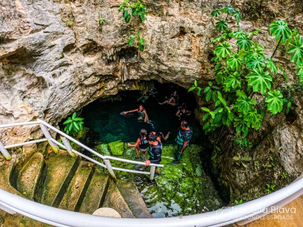 A group of people explore a natural cave pool surrounded by rocky walls and lush greenery, accessed by stone stairs with a railing.