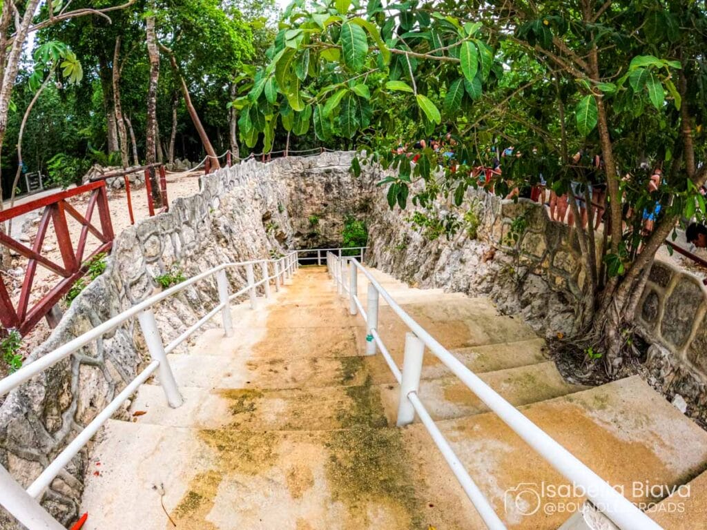 Stone staircase descends into a cenote, surrounded by lush vegetation. People observe from a wooden railing. The scene feels serene and inviting.