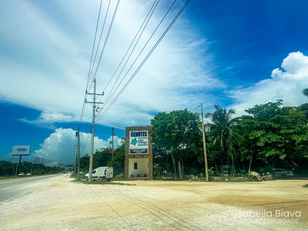 An open road under a bright blue sky with clouds and a sign for Cenotes Casa Tortuga. Lush greenery lines the roadside.