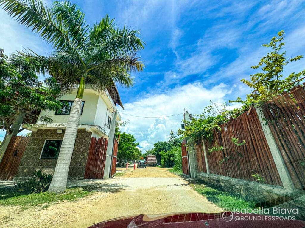 A two-story building with palm trees and a wooden fence under a vibrant blue sky on a dirt road, possibly near a construction site.