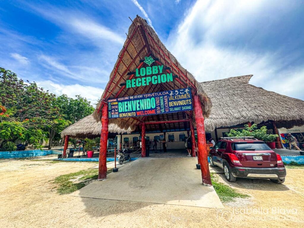 Thatched-roof reception area with colorful multilingual welcome signs. Red SUV parked nearby. Vibrant greenery surrounds the structure. Blue sky with scattered clouds above.