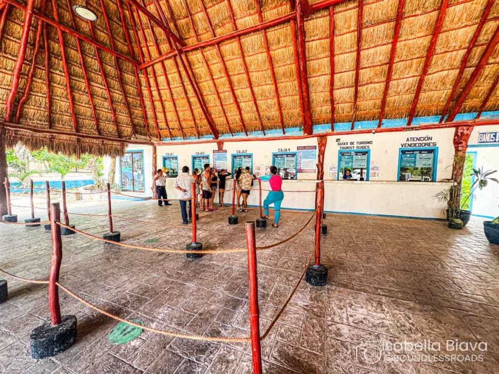 A group of people is gathered in a thatched-roof building, queuing at a tourist information and ticket counter.