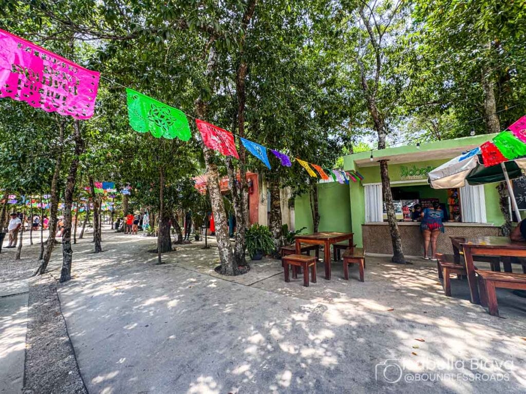 Outdoor area with colorful flags, tables, and a small cafe surrounded by trees. A person stands near the counter.