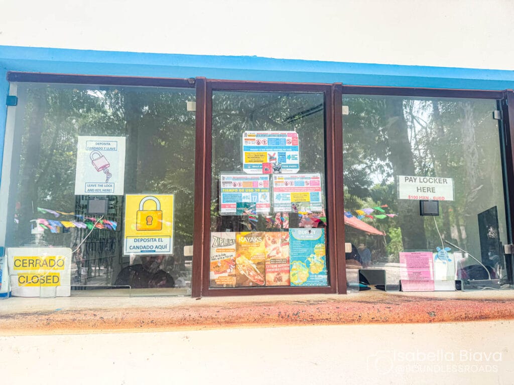 A small ticket window with colorful notices and signs. Trees and decorative flags are visible in the reflection. No recognizable landmarks present.