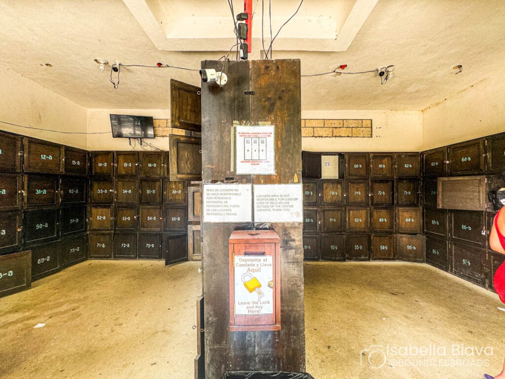 Room with numerous dark wooden lockers, some open. Overhead wires and signs posted on doors. Person partially visible in the corner.