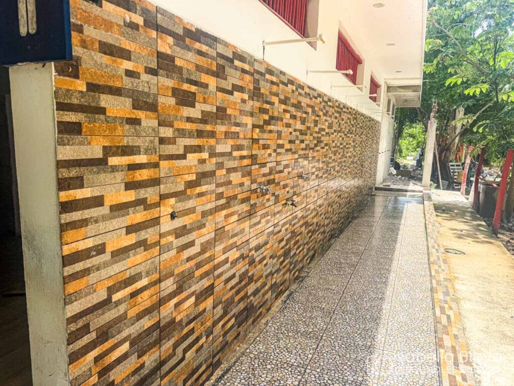 A tiled wall and walkway beside a building with red window accents, surrounded by trees under clear sunlight.
