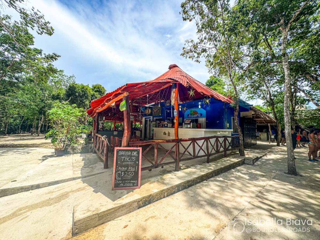 A colorful outdoor taco bar with a red roof, surrounded by trees. People gather nearby under the sunny sky.